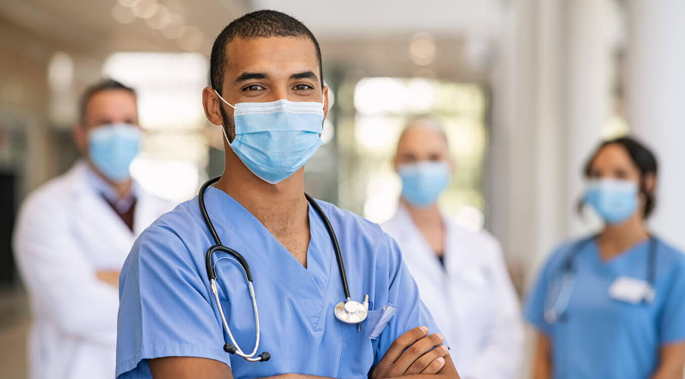 Foco em enfermeiro com roupa azul e máscara. Ao fundo, desfocado, há dois médicos e uma enfermeira 