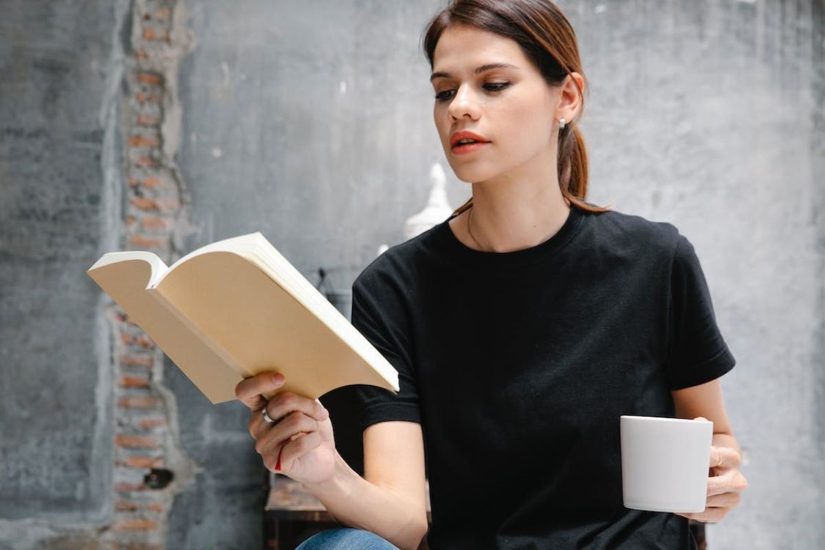 Uma pessoa está segurando uma xícara na mão e um livro no outro. Ela está de camisa preta e na frente de uma parede cinza