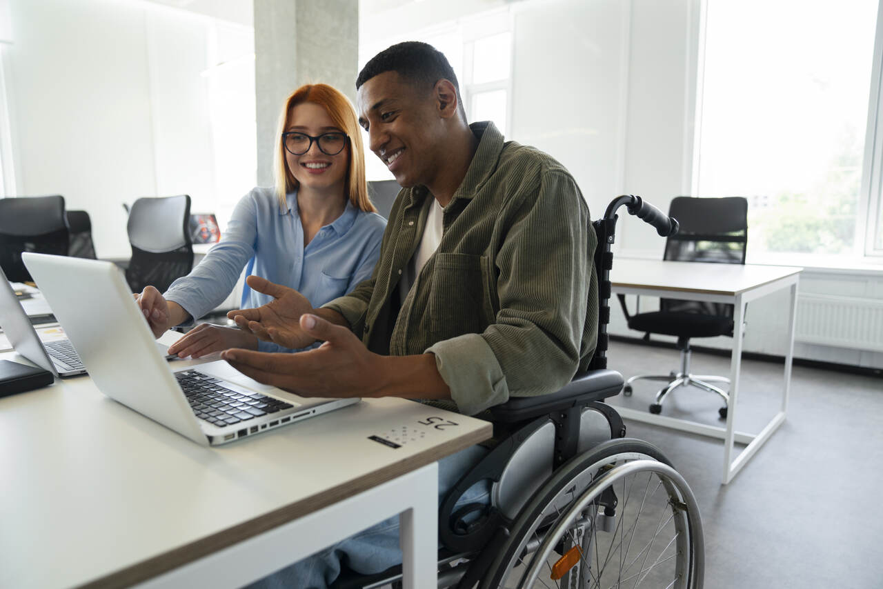 jovem cadeirante discute sobre algo na tela de seu notebook com colega de trabalho sentada ao seu lado