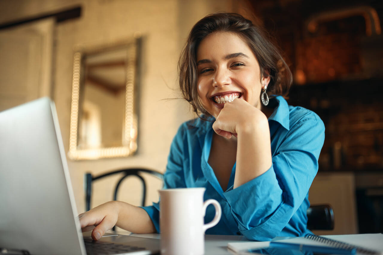 Jovem posa sorridente enquanto faz algo em seu notebook