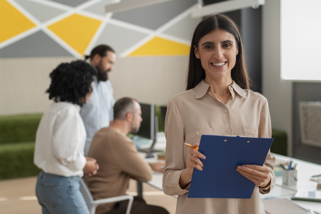 Profissional de RH possa sorridente segurando uma prancheta com colegas de empresa ao fundo 